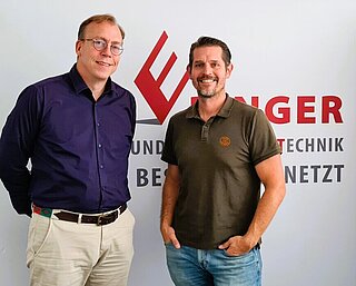 Picture shows two men in front of a billboard of the company NST Ebinger GmbH Salzburg