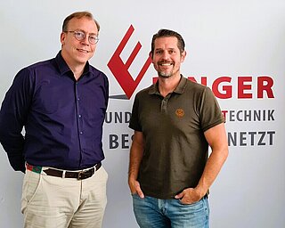 Picture shows two men in front of a billboard of the company NST Ebinger GmbH Salzburg