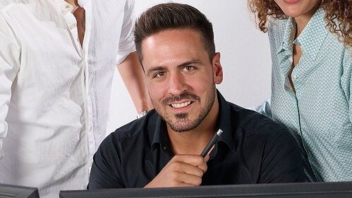 Man smiling sitting behind laptop with pen in hand .